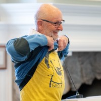 Man lifts up sweater to show yellow t-shirt underneath that says G.V.S.C.